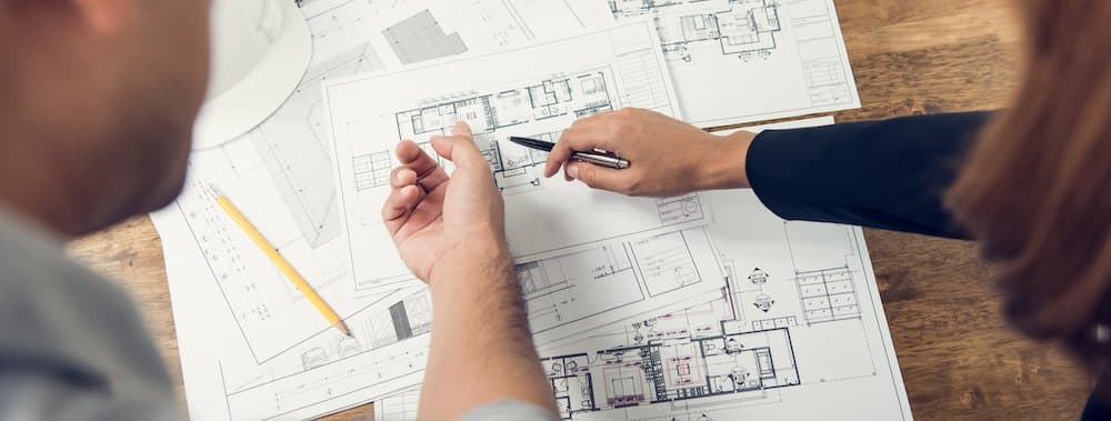 Two people point to different rooms in a blueprint of a floor plan on the wooden table in front of them.