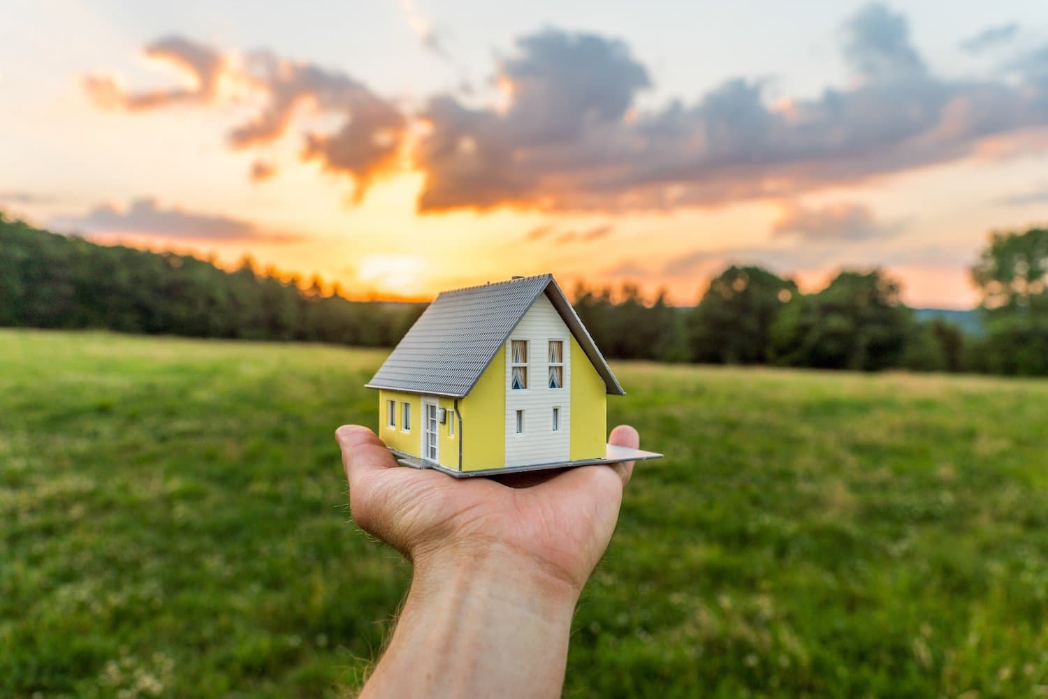 A hand holds up a small yellow model home in their palm. In the background is a field lined with trees. The sun is setti