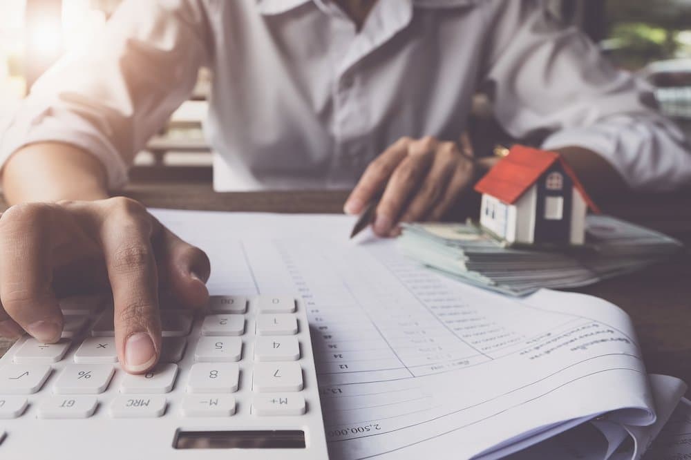 A person sits at a desk with a small model home and money next to them to illustrate the costs of buying a home. They us