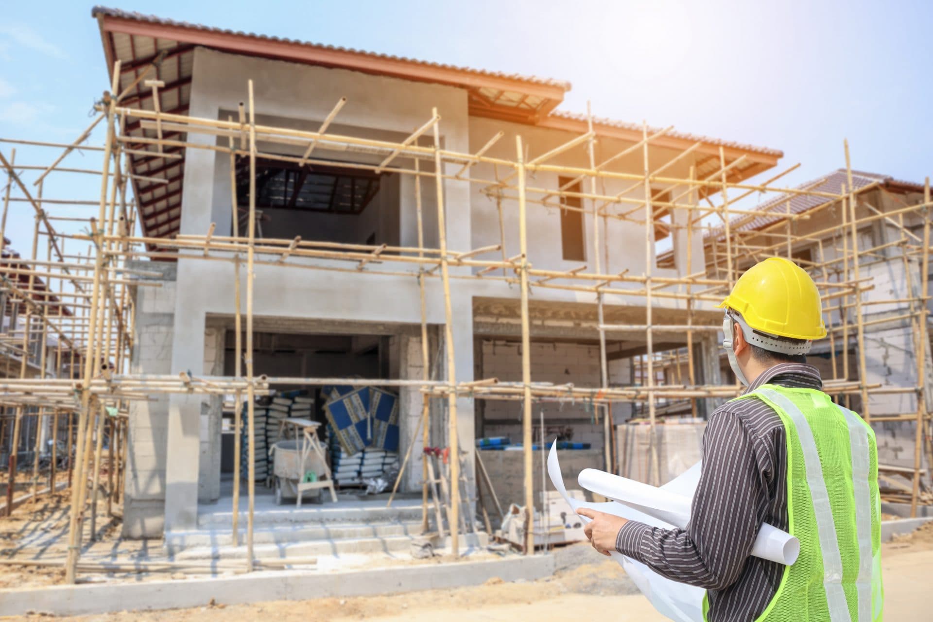 An engineer in a hard hat and hi-vis stands with blueprints looking at a home under construction, surrounded by scaffold